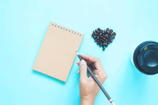 Diseño plano creativo de granos de café en forma de corazón y lápiz de mano de mujer con cuaderno sobre fondo de color azul — Foto de Stock