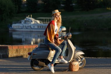 Çekici bir kızın portresi. Kot pantolonlu kadın, kısa tişört ve hasır şapkalı kadın gün batımında nehrin kenarında eski bir motosikletin üzerinde oturuyor. Seyahat kavramı.