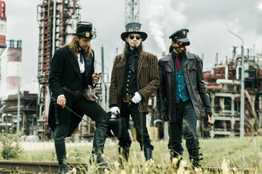 Steampunk tarzı üç adam endüstriyel arka planda duruyor.