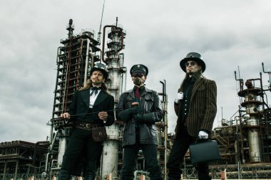 Steampunk tarzında üç adam endüstriyel arka planda ayakta durmayı tartışıyor.