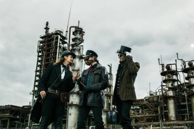 Steampunk tarzında üç adam endüstriyel arka planda ayakta durmayı tartışıyor.