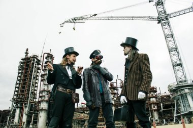 Steampunk tarzında üç adam endüstriyel arka planda ayakta durmayı tartışıyor.