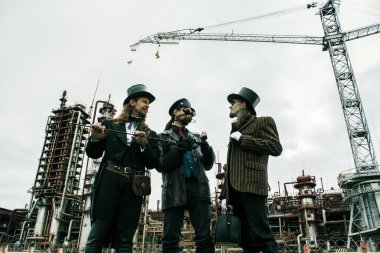 Steampunk tarzında üç adam endüstriyel arka planda ayakta durmayı tartışıyor.