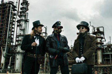 Steampunk tarzında üç adam endüstriyel arka planda ayakta durmayı tartışıyor.