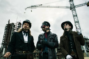 Steampunk tarzında üç adam endüstriyel arka planda ayakta durmayı tartışıyor.