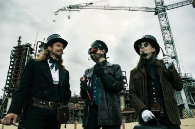 Steampunk tarzında üç adam endüstriyel arka planda ayakta durmayı tartışıyor.