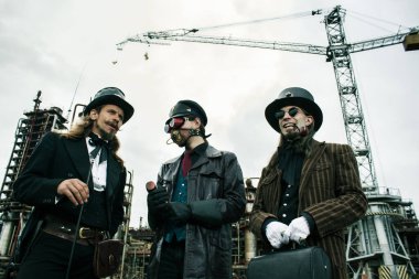 Steampunk tarzında üç adam endüstriyel arka planda ayakta durmayı tartışıyor.