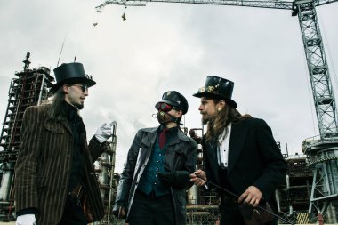 Steampunk tarzında üç adam endüstriyel arka planda ayakta durmayı tartışıyor.