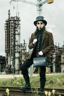 Steampunk tarzı giyinmiş genç adam endüstriyel arka planda poz veriyor.