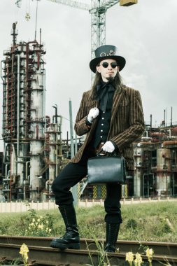 Steampunk tarzı giyinmiş genç adam endüstriyel arka planda poz veriyor.