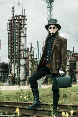 Steampunk tarzı giyinmiş genç adam endüstriyel arka planda poz veriyor.