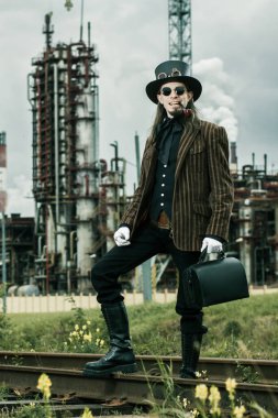 Steampunk tarzı giyinmiş genç adam endüstriyel arka planda poz veriyor.