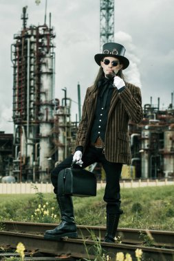 Steampunk tarzı giyinmiş genç adam endüstriyel arka planda poz veriyor.