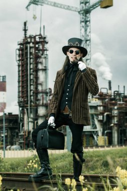 Steampunk tarzı giyinmiş genç adam endüstriyel arka planda poz veriyor.