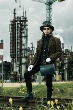 Steampunk tarzı giyinmiş genç adam endüstriyel arka planda poz veriyor.