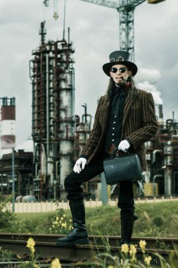 Steampunk tarzı giyinmiş genç adam endüstriyel arka planda poz veriyor.