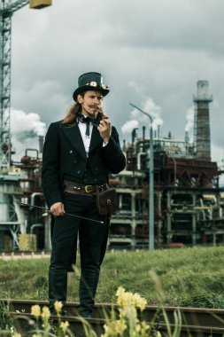 Steampunk tarzı giyinmiş genç adam endüstriyel arka planda poz veriyor.