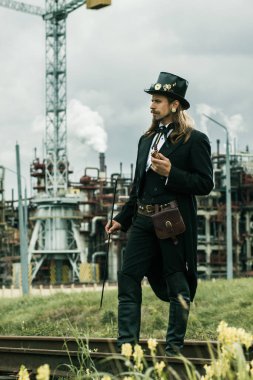 Steampunk tarzı giyinmiş genç adam endüstriyel arka planda poz veriyor.
