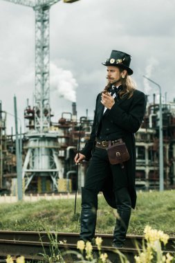 Steampunk tarzı giyinmiş genç adam endüstriyel arka planda poz veriyor.