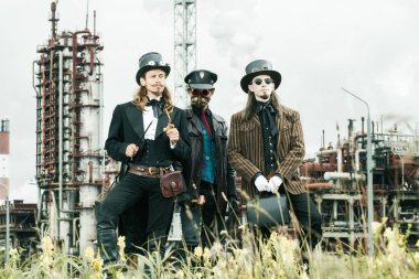 Steampunk tarzı üç adam endüstriyel arka planda duruyor.