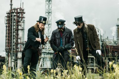 Steampunk tarzı üç adam endüstriyel arka planda duruyor.
