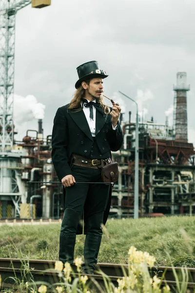 Steampunk tarzı giyinmiş genç adam endüstriyel arka planda poz veriyor.
