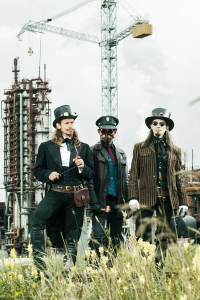 Steampunk tarzı üç adam endüstriyel arka planda duruyor.