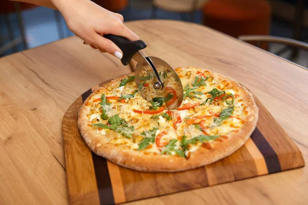 Female hand with circle knife cutting freshly pizza slices on wooden board in kitchen, Soft focus. Hand slicing pizza with cutter on table.