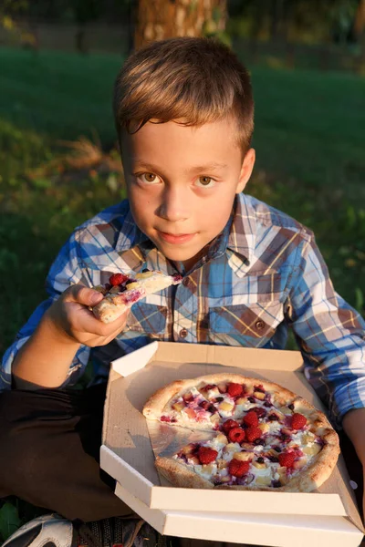 The boy eat a piece of pizza. Picnic on the grass. Boy have a dinner with sweet pizza on the meadow .