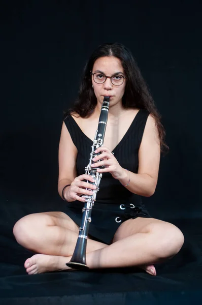 A brunette girl with her clarinet, with glasses, lying down laughing. on a black uniform background