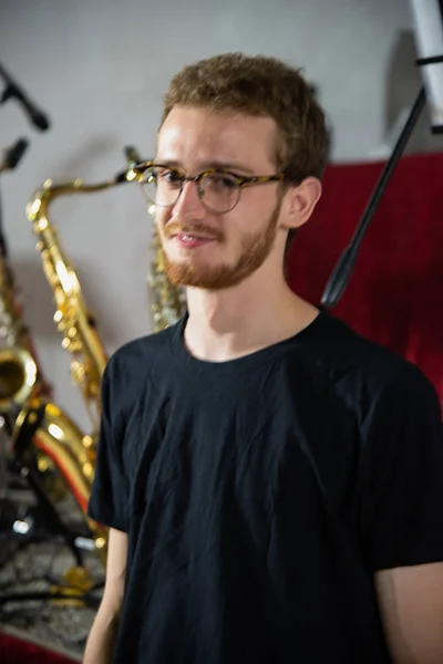 Young handsome tall guy posing in black t-shirt, musical instruments background, boy with glasses and beard, young musician portrait before a concert