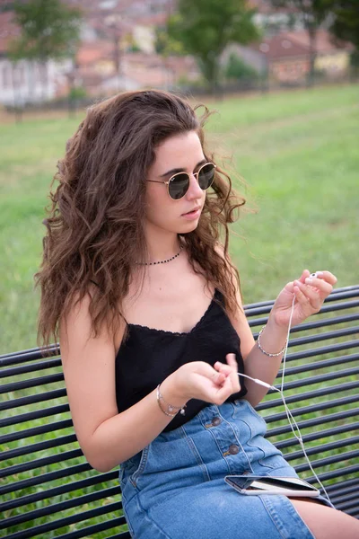 Young beautiful girl sitting on park bench with headphones listening to music on smartphone outdoors. Young girl with long brown hair with sunglasses relaxed