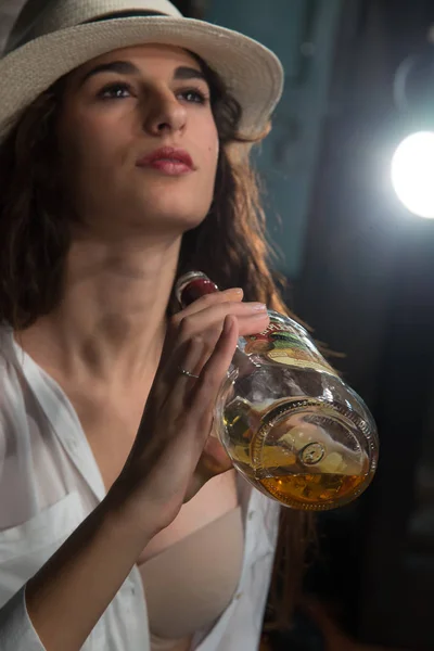 Young girl with wavy brown hair and white hat on her head. With a bottle of whiskey in his hand. Holds open the white shirt, flesh-colored bra, very sensual. Light in the background. Look lost in the air. Sensuality concept of young generations.
