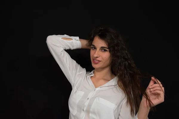 Young beautiful girl with long wavy brown hair and arms in her hair. He wears a white men's shirt, a smiling and sensual attitude. Half-length portrait. Black background. Copy space