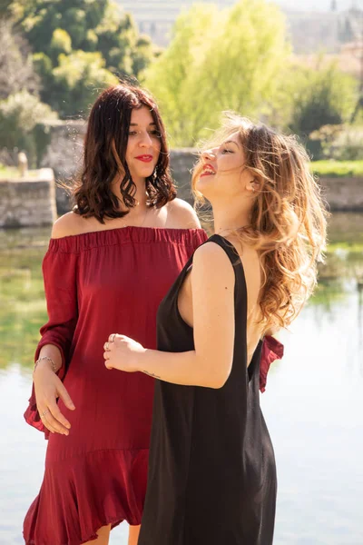 Two female friends happily dance outdoors along the shores of a lake. A blonde and black dress, the other brunette in a red dress. Youth fun concept.