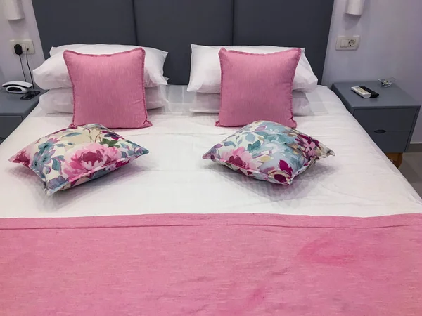 Double bed with white and pink sheets. Hotel bedroom with remote controls and colorful pillows.