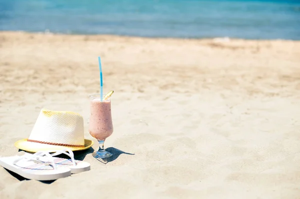 Клубничный свежий фруктовый сок, шляпа и тапочки возле моря на песчаном пляже — стоковое фото
