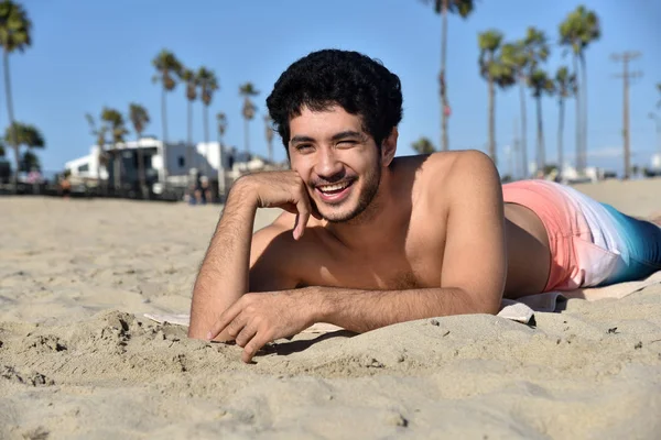 A handsome young man laying in the sand on the beach with a big smile
