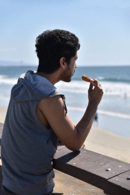 Yakışıklı genç adam Pasifik Okyanusu'na bakan iskelede bir dilim pizza yeme