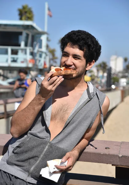 Happy young man eating a slice of pizza on the pier near the lifeguard hut