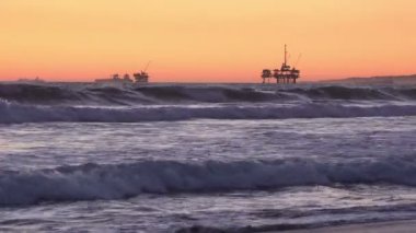 Dalgalar offshore petrol sondaj kuleleri ile gün batımında kıyıya çöküyor