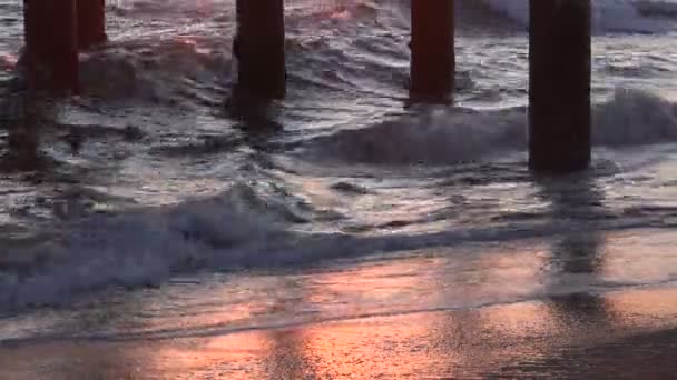 夕日によって強調された桟橋の下で岸に波が墜落する — ストック動画