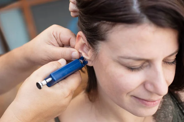 Woman having ear piercing process with special piercing gun in beauty center by medical worker, cropped close up view