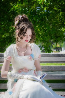 young girl in a smart long white dress is reading a love story on a park bench and is enjoying her. clipart