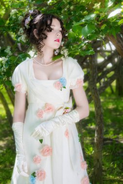 Zarif, uzun, beyaz gelinlikli genç kız bir bahar bahçesinde çiçek açan çalıların yanında..