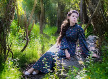 güzel genç üzgün kadın Boho tarzda giyinmiş, bir orman içinde bir ağaç tarafından bir taşa oturur ve üzüntü ile mesafede görünüyor.