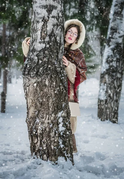 ロシアの女の子の明るいナショナルのスカーフ 毛皮のコートを寒い冬の日に皮を再生 雪に覆われた公園の古いバーチの後ろから外を見てかくれんぼ — ストック写真