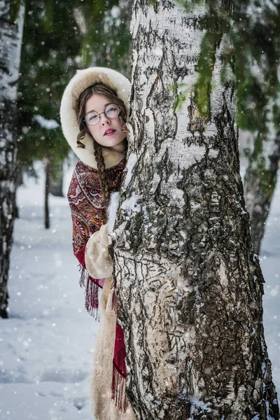 ロシアの女の子の明るいナショナルのスカーフ 毛皮のコートを寒い冬の日に皮を再生 雪に覆われた公園の古いバーチの後ろから外を見てかくれんぼ — ストック写真