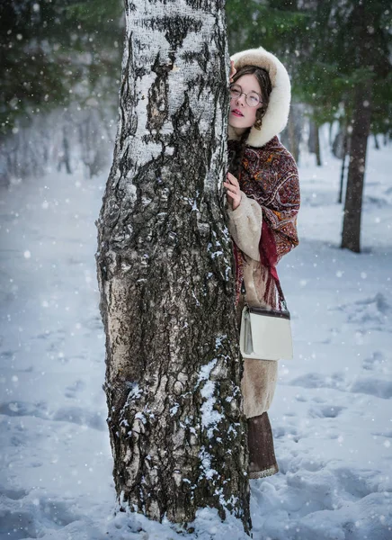 ロシアの女の子の明るいナショナルのスカーフ 毛皮のコートを寒い冬の日に皮を再生 雪に覆われた公園の古いバーチの後ろから外を見てかくれんぼ — ストック写真