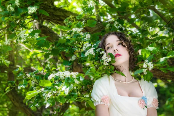 春の庭に咲く木の近くにエレガントな長い白い花嫁のドレスの若い女性 — ストック写真